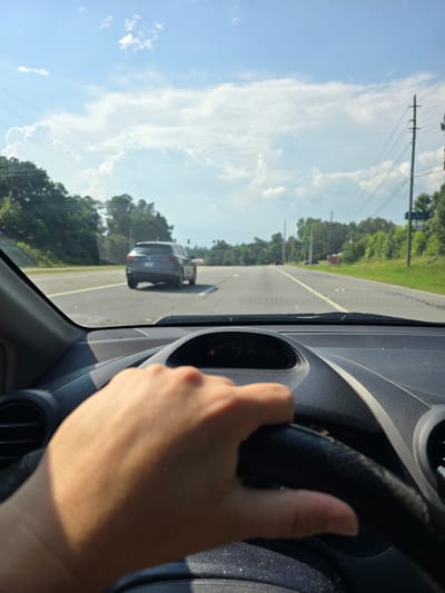 A first-person perspective shot from inside a vehicle shows a hand holding a steering wheel while driving on a sunny day. The vehicle is traveling on a paved road with multiple lanes and a white dividing line. A dark gray SUV is visible in front of the vehicle, also driving on the road. The surrounding environment consists of lush green trees lining the sides of the road, power lines supported by utility poles, and a bright blue sky with scattered white clouds. The dashboard of the car is visible, including a digital display showing "47" which likely indicates the speed in miles per hour. There is no indication of the location being specifically within the city of Woodstock, United States, based solely on the visual information provided in the image.