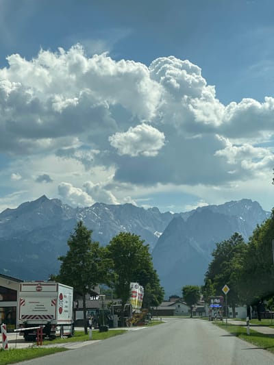 The image depicts a street scene in Farchant, Germany, with a backdrop of the Zugspitze mountain range. In the foreground, a white truck with red and white striped markings on the rear is parked on the right side of the road. The truck has text in German on its side, including "VERKEHRS- TECHNIK SICHERUNG PLANUNGSBÜRO". A white barrier with red and white stripes is in front of the truck.
To the left of the truck, there is a building with a gray facade and a brown roof. Further down the street, to the right, a diamond-shaped yellow road sign with a black border and a black symbol is visible. Adjacent to this sign is a red circular sign with a white horizontal bar. Behind these signs, there are buildings with tiled roofs and trees.
The middle ground is dominated by a line of lush green trees. Beyond the trees, the majestic Zugspitze mountain range rises, with its peaks partially covered in snow. The mountains are sharp and rugged, with visible ridges and valleys.
The sky is a vibrant blue, with large, dramatic white cumulus clouds, some of which appear to be forming thunderheads. The sunlight casts a bright light on the scene, creating strong contrasts. The overall impression is a scenic view of a road leading towards the mountains on a cloudy but bright day.