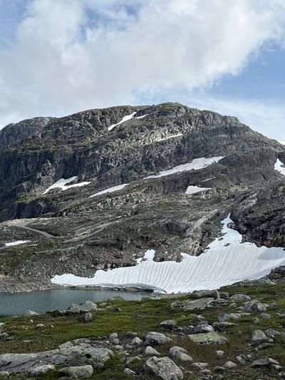 The image depicts a mountainous landscape with a body of water in the foreground. The mountain is rocky and steep, with patches of snow scattered across its slopes. A winding road is visible ascending the mountainside. The foreground consists of green vegetation and scattered rocks. The sky is partly cloudy. This scene is located in Brattabø, Norway.