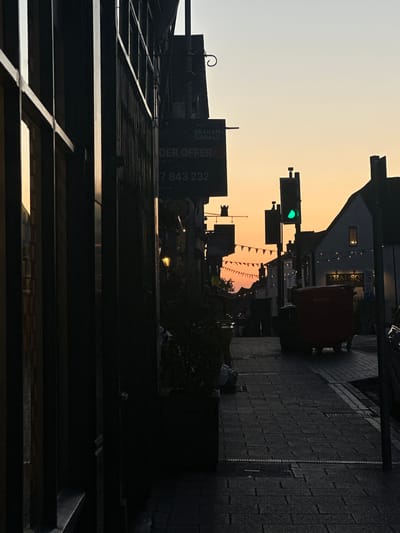 The image depicts a street scene in St Albans, United Kingdom, at dusk or dawn. The view is looking down a street lined with buildings. A traffic light is illuminated with a green light. Pennant flags are strung across the street. A sign for "Graham Sibbald" is visible on a building. The sky is a gradient of yellow and light grey. The street is paved with cobblestones.