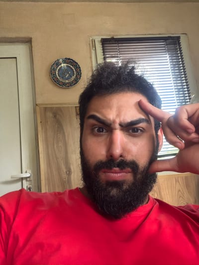 A close-up, upright portrait captures a man indoors, looking directly at the camera. He has dark, curly hair and a full beard, and is wearing a plain red t-shirt. His expression is intense, with a furrowed brow and slightly pursed lips. His right hand is raised, with the index finger touching his temple and the thumb pointing downwards, forming a gesture often associated with thought or an "L" shape.
The setting is a room with light-colored, possibly beige, walls. On the left side of the frame, a window with vertical blinds allows bright daylight to enter, suggesting it is daytime. Below the window, a decorative, circular plate is affixed to the wall. This plate is intricately patterned in blue, red, and white, and features a central geometric motif that resembles a compass and square. Visible text curved around the central motif includes "MÜHENDİS" and "YAPI". To the right, a wooden surface, possibly a cabinet or wall panel, and a white door or frame with a simple white handle are discernible.