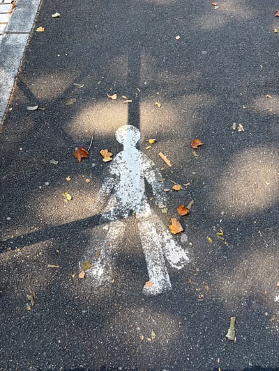 The image displays a faded white pedestrian crossing marking on a dark asphalt surface, likely a path or road. The marking depicts a stylized human figure with outstretched arms and legs, suggesting movement. Scattered around the marking are several fallen autumn leaves in shades of brown and yellow, along with a few small twigs. The scene is partially illuminated by sunlight filtering through unseen foliage, casting dappled shadows across the asphalt. The background includes a grey paved area with white lines on the left edge of the frame, possibly a sidewalk or border. The overall impression is one of an outdoor public space in Charnay-lès-Mâcon, France, during the autumn season.