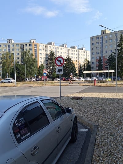 The image shows a street scene in Zvolen, Slovakia. In the foreground, a silver car is parked on a gravel lot next to a paved road. The car has several stickers on its rear window, including one that reads "GIRL RACER." The road is lined with trees and buildings, including several apartment blocks. Traffic signs are visible, including a sign indicating a left turn is prohibited and another showing a pedestrian crossing. A red fire hydrant stands near the sidewalk. The sky is blue with some clouds, suggesting a clear day. The overall scene appears to be a residential or mixed-use urban area. The presence of parked cars, apartment buildings, and street infrastructure points to a daytime setting, likely during business hours or late afternoon. The car stickers suggest a car enthusiast or custom car culture element.