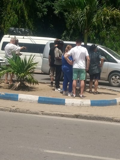This image depicts an outdoor scene in the Ganjoni/Shimanzi ward of Kenya, under bright sunlight. Seven individuals are visible near two parked vehicles on what appears to be a roadside or parking area adjacent to a paved road.
To the left, a white van, possibly a Toyota Hiace, is parked with its rear facing partially towards the camera. It has a roof rack loaded with multiple black bags. A partial license plate reading "KCD 213" is visible. Two males stand near this van; one man in a white short-sleeved shirt gestures with his right arm towards the left, while another individual, partially obscured, stands nearby.
To the right of the scene, clustered around the front of a silver minivan or SUV (partial front right side visible), are five individuals. From left to right within this group, there is a male in a black t-shirt and light brown shorts; a male in a striped t-shirt and dark trousers; a female in a blue jumpsuit or dress with white sneakers; a male in a white t-shirt and light green shorts; and a final individual (gender indeterminate from this angle, possibly female) wearing a dark patterned top, dark shorts, and a hat, with hands on hips. This group appears to be standing and observing or conversing, facing generally towards the right and away from the camera.
The ground where the people are standing is a dry, earthy patch. A small, green palm-like plant grows in this area, near a curb. The curb is painted with alternating blue and white sections, separating the dirt area from a grey asphalt road that occupies the foreground of the image. Lush green foliage and trees form the background.