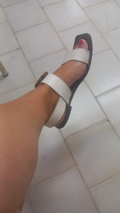 The image shows a close-up of a person's left foot and lower leg, wearing a white sandal with a dark sole and a brown buckle. The sandal has a square toe. The foot is positioned on a tiled floor, which appears to be made of light-colored square tiles with dark grout lines. The tiles are arranged in a grid pattern. The background is out of focus and shows more of the tiled floor. There is a faint drawing of a "D" on one of the tiles. The lighting suggests an indoor setting. The image appears to be taken from a slightly elevated angle, looking down at the foot. The overall impression is that of a casual snapshot. The location is Águeda, Portugal, though no specific landmarks are visible to confirm this.
