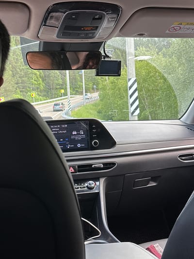 This image shows the interior of a car, looking forward through the windshield. The car is traveling on a road with trees and greenery on either side. Yellow road signs are visible along the road. The dashboard of the car is visible, including the infotainment screen which displays "Sat, Jun. 20 16:28" and "USB Music" with a song title "ejima - Life.mp3". The car is likely in motion, as indicated by the road and scenery passing by. The image does not provide information about the specific location of Bol'shaya Rechka, Russia.