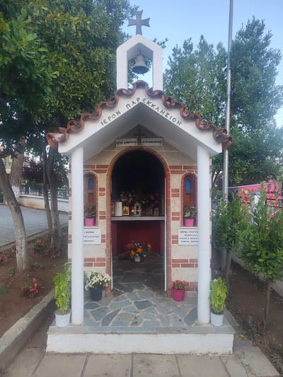 This media file shows an exterior view of a small, traditional Greek Orthodox chapel, possibly a proskynetarion, located in Νέα Ραιδεστός, Greece. The chapel is adorned with a red-tiled, wavy roof and a white bell tower featuring a cross. Inscriptions in Greek are visible on the chapel's facade. Above the entrance, the text reads "ΙΕΡΟΝ ΠΑΡΕΚΚΛΗΣΙON," meaning "Holy Chapel." To the left and right of the entrance, smaller plaques identify saints: "ΑΓΙΟΣ ΔΗΜΗΤΡΙΟΣ" (Saint Demetrius) and "ΑΓΙΟΣ ΚΩΝΣΤΑΝΤΙΝΟΣ" (Saint Constantine). Below the entrance, more inscriptions are present, including "Σ ΜΝΗΜΗΝ" (In memory of) and names that appear to be family names. Another inscription on the right side reads "ΔΩΡΕΑ ΟΙΚΟΓΕΝ. ΜΑΝΩΛΑΚΗ" (Gift of the Manolakis family), followed by a list of names.
The chapel is situated outdoors, with trees and greenery surrounding it. Several potted plants, including flowers and a small green shrub, are placed around the entrance, adding color and life to the scene. The chapel is built with stone and brick accents, and the pathway leading to it is paved with irregular stones. The time of day appears to be late afternoon or early evening, as indicated by the soft, diffused lighting. The weather seems pleasant, with clear skies visible. The overall impression is one of peace and devotion, characteristic of a religious sanctuary in a community setting.