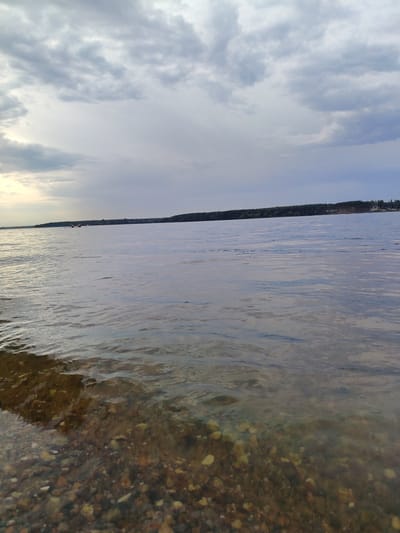 The image depicts a waterfront scene at the Tourist Resort "Klyazma Reservoir" in Russia. The foreground features a shallow body of water with a rocky, pebble-strewn bottom visible through the clear water. The water surface exhibits gentle ripples. In the mid-ground, the expansive Klyazma Reservoir stretches out, reflecting the overcast sky. A distant shoreline, lined with a dense forest, forms the horizon. A few sailboats are visible on the water in the distance. The sky is filled with a mixture of light and dark clouds, suggesting an overcast day. The overall atmosphere is calm and serene, characteristic of a natural landscape.
