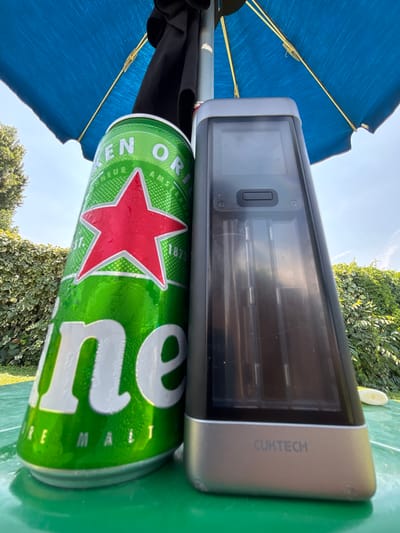 The image displays a close-up, rotated view of a Heineken beer can and a silver Cuktech device resting on a green surface, likely a table. The background suggests an outdoor setting with lush green foliage and a bright sky, indicating it is daytime with clear weather. A portion of a blue umbrella is visible on the left side of the frame, hinting at sun protection.
The Heineken can, a popular brand, is predominantly green with its iconic red star and white lettering. The Cuktech device, a piece of technology, has a silver casing and a dark screen displaying internal components. Text visible on the device reads "CUKTECH". The overall impression is a relaxed, casual outdoor moment, perhaps during a break or a leisurely activity in a garden or patio area. The presence of the beer and the device could suggest someone is enjoying a drink while using or testing the technology outdoors. The lighting suggests it's midday, with the sun casting shadows. There are no people visible in the image.