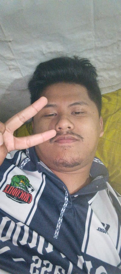 A young man is lying down and taking a selfie. He is wearing a blue and white striped shirt with a logo of a crocodile on the left chest. He has short black hair and a mustache and goatee. He is holding up two fingers in a peace sign. The background is a light-colored, textured surface, possibly a bedsheet. The image is grainy and the lighting is dim. The location is stated to be Kemuning, Indonesia.