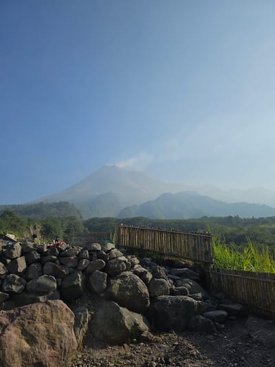 The image depicts a volcanic landscape under a clear blue sky. In the background, a large volcano, likely Mount Merapi, is visible, emitting a plume of smoke from its summit. The slopes of the volcano and surrounding mountains are covered in lush green vegetation, with layers of mist or haze obscuring the distant peaks.

In the foreground, a path or viewing area is constructed with large rocks and gravel. A rustic bamboo fence runs along the right side of the path, separating it from a dense area of tall green grass. To the left of the path, a substantial pile of large, weathered rocks forms a natural barrier. Among these rocks, a red ATV-like vehicle is parked, and two individuals are visible near it. One person appears to be standing by the vehicle, while another is further back, possibly near a set of concrete steps.

The scene suggests an outdoor recreational area or a viewpoint overlooking the volcano, likely located in or near Cangkringan, Indonesia, a region known for its proximity to Mount Merapi and its tourism activities. The lighting indicates it is daytime, with the sun casting shadows and illuminating the landscape.