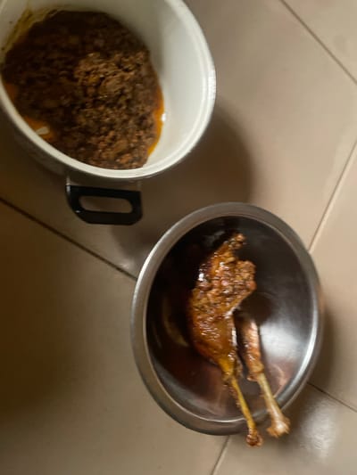The image shows two metal bowls on a tiled floor. One bowl, larger and white on the outside, contains a dark, textured dish. The other, smaller and metallic, contains two pieces of cooked meat with visible bones. The tiles are a neutral beige color, and the lighting is dim. The scene appears to be indoors. No people are visible. The image does not provide any information that would allow for the localization of Aiyegunle, Nigeria.