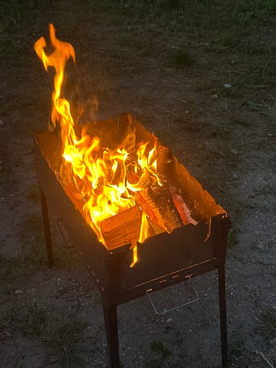 A top-down, slightly angled view of a fire burning in a metal brazier on a dark, grainy ground, with patches of grass. The fire is vibrant with bright orange and yellow flames consuming several pieces of wood, one of which is a large, light-brown log positioned in the upper right of the brazier. The brazier is dark and rectangular with a metal handle on the right side. The fire is actively burning, with smoke trailing to the left. The ground is composed of dirt, small stones, and some dry grass. The lighting suggests it is dusk or evening, with no clear weather indicators. The scene evokes a feeling of warmth and outdoor activity, likely a campfire or barbecue. No people or text are visible. The location context provided is Velyka Cherniavka, Ukraine.
