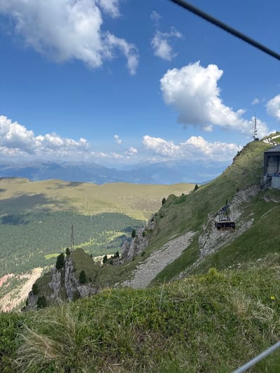 This image captures a breathtaking mountainous landscape. The scene is dominated by a steep, grassy mountainside that plunges into a valley dotted with trees. In the distance, layers of more mountains fade into a hazy blue. The sky is a vibrant blue, punctuated by fluffy white clouds.
A cable car is suspended on a wire, its carriage clearly visible. The carriage has "SECEDA" written in yellow letters on a black background. At the top of the mountain, near the cable car station, a building structure is visible, along with a telecommunications tower. The time of day appears to be daytime, given the bright sunlight illuminating the scene. The weather seems pleasant with clear skies and clouds. The presence of the cable car and the building suggests this is a popular tourist destination, likely a ski resort or hiking area in the Alps, given the geographical context provided. The overall impression is one of natural beauty and recreational activity in a high-altitude setting.