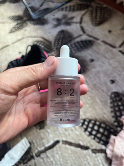 This is a close-up, eye-level shot of a person's hand holding a small, clear bottle of "Perfect Balance Facial Serum" by B:Lab. The bottle has a white dropper cap and contains a light purple liquid. The label on the bottle is partially obscured by the hand but is readable in parts. Visible text includes "Perfect Balance Facial Serum," the ratio "8:2," ingredients like "Erythrorhizon" and "Aloe vera," manufacturing details, usage instructions, and the brand name "B:Lab." The background is out of focus, showing a patterned rug or carpet in muted colors, with hints of pink and black fabric visible. The lighting suggests an indoor setting, possibly during the daytime. The image captures a moment of showcasing a skincare product.