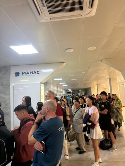 The image shows a queue of people in an airport terminal. A large sign with the MAHCAC logo and text in Cyrillic characters is visible on the left side of the image, indicating the location is Manas International Airport in Bishkek, Kyrgyzstan. The ceiling features fluorescent lighting and an air conditioning unit. The people in the queue are diverse in age and gender, dressed in casual clothing, and some are holding luggage. They appear to be waiting in line, possibly for check-in or security. The environment is indoors, with tiled floors and plain walls.