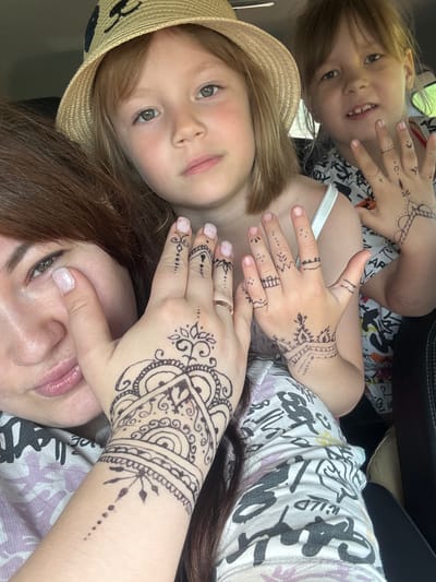 The image depicts three individuals, likely a mother and her two children, posing for a selfie indoors, possibly in a car given the dark interior visible. The woman in the foreground is on the left, showing her face and hand adorned with intricate henna tattoos. She has long, dark brown hair and is wearing a white top with a patterned design. She is touching her face with her left hand, which is prominently displayed with henna. To her right are two children. The older child, in the middle, is wearing a straw hat with a dark design and looking directly at the camera. The youngest child is in the background, to the right, also looking towards the camera and displaying henna on their hand. The lighting suggests it is daytime, and the overall impression is one of shared activity and enjoyment. The location in the city of Аряж, Russia is not discernible from the image itself.