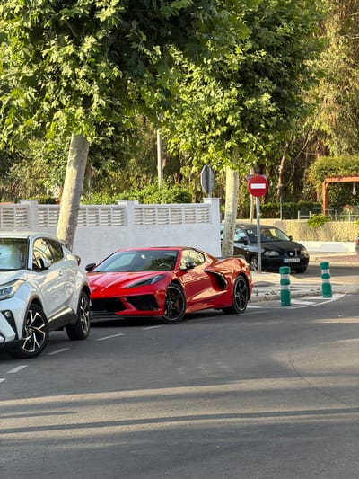 The image depicts a street scene in Pla Parcial Platja, Spain. In the foreground, a red Chevrolet Corvette C8 is parked, facing right. To its left, a white Toyota C-HR is parked, facing left, with its front portion visible. Behind these vehicles, a black SEAT Ibiza is parked, facing right, displaying a license plate that reads "0340 CYS". The cars are parked on an asphalt road with white lane markings. A white, slatted wall runs along the background, with trees and foliage behind it. A red circular traffic sign with a white horizontal bar, indicating "no entry", is visible on a pole to the right of the white wall. Two green and white bollards are positioned on the sidewalk next to the street. The lighting suggests it is late afternoon or early evening, with long shadows cast across the road. The environment appears to be a residential or urban area with vegetation.