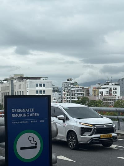 An outdoor scene captured under a cloudy, overcast sky depicts an urban environment in Hòa Cường Ward, Da Nang, Vietnam. In the foreground, a dark blue rectangular sign, partially cropped on the left, displays "DESIGNATED SMOKING AREA" in English, Vietnamese ("KHU VỰC HÚT THUỐC"), Korean, and Chinese/Japanese scripts. Below the text, a white circular icon with a green border features a stylized lit cigarette emitting smoke.

In the midground, a white Mitsubishi Xpander minivan is positioned on a paved road, facing slightly left. The vehicle's front grille features the Mitsubishi emblem, and its license plate reads "43H-240.82." A red "TP" logo is visible on the front passenger door. A gray metal barrier separates the sign from the road and continues horizontally across the frame. Sparse red foliage is visible behind the barrier to the right of the sign.

The background reveals a dense urban skyline composed of various multi-story buildings, predominantly in shades of white, beige, and gray. Some buildings feature visible windows, balconies, and rooftop antennae or structures. Green trees are interspersed throughout the urban landscape. Distant, hazy mountains or hills are visible beneath the low, gray clouds on the horizon. No individuals are present in the image.