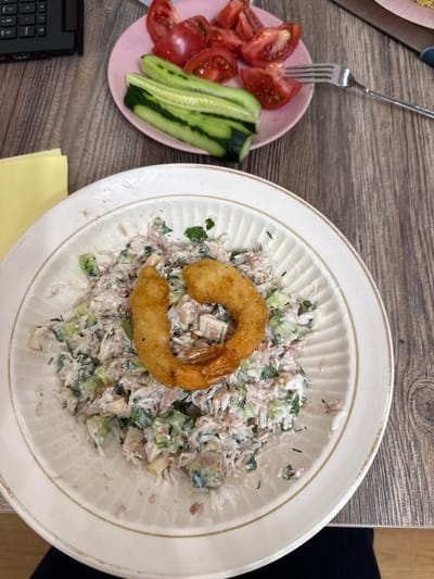 A top-down view shows a meal on a wooden table, with a plate of salad and a side of sliced tomatoes and cucumber. The salad, served on a fluted white plate with a gold rim, appears to be a tuna or chicken salad mixed with chopped vegetables and herbs, possibly with rice. A single, large, golden-brown fried shrimp is placed on top of the salad, curved into a semi-circle.

To the left of the main plate, a smaller pink saucer holds sliced red tomatoes and strips of green cucumber. A silver fork rests on the table, its tines partially inserted into the pink saucer. The wooden table surface shows a visible grain pattern. In the bottom left corner, a black laptop keyboard is visible, suggesting a casual or work-from-home setting. A yellow sticky note is also partially visible on the table.

The lighting suggests it might be daytime, with diffused light. There are no people visible in the frame. The scene is focused on the food, arranged for consumption. The overall impression is of a prepared meal, ready to be eaten.