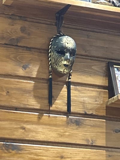An ornate, dark-colored mask with gold accents hangs against a light brown, horizontally paneled wooden wall in Bukovlak, Bulgaria. The mask, appearing to be a decorative Venetian-style piece, features intricate gold scrollwork on the forehead and right cheek, with a gold scale pattern on the left cheek. Braided gold and black cords descend from each side of the mask, terminating in black tassels. The mask is suspended from a dark, L-shaped bracket attached to the underside of a light-colored wooden shelf running horizontally across the top of the frame. To the right, a smaller wooden shelf is partially visible, holding a dark-framed object, likely a picture or mirror. The wooden paneling exhibits natural grain and knots. No individuals are present, and no actions or interactions are depicted.