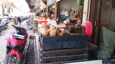 The image depicts a market stall in Puloara, Indonesia, with a focus on a display of coconuts. A red scooter is parked prominently in the foreground to the left, its license plate reading "BL 3623 DBH 10-27". Behind the scooter, a row of parked motorcycles lines a paved pathway. The stall itself features a large wooden crate filled with numerous coconuts. To the right of the coconuts, a motor and a large pink basin are visible. In the background, several individuals can be seen engaged in market activities. One person in a red and white shirt is attending to goods behind a counter, and another person in white is also present. The stall is open to the street, with awnings providing shade overhead. Bags of goods and other market items are also visible, suggesting a typical bustling marketplace environment.