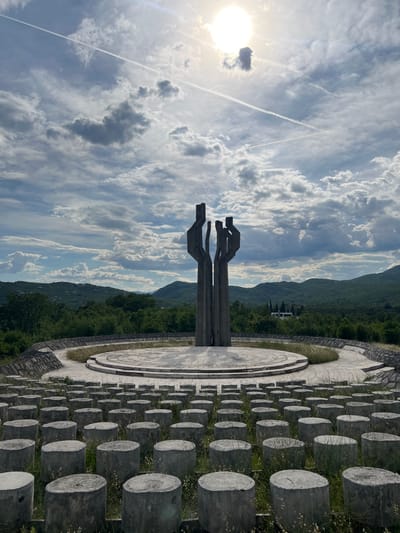 The image depicts the Barutana Memorial Complex in Montenegro. A prominent concrete monument stands against a backdrop of a cloudy sky, mountains, and dense green forest. The monument is a sculpture consisting of three vertical elements, each with a distinct horizontal extension at the top, appearing like abstract figures or hands. At the base of the monument is a circular, paved area. Surrounding this area is a field with a grid of gray concrete cylinders, possibly representing graves or individuals. The overall environment is serene, yet carries a somber tone.