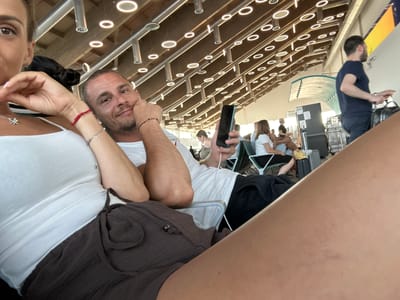 This is a selfie taken by a couple in what appears to be an airport waiting area. The image is oriented sideways. The man, in the foreground, is looking towards the camera with a slight smile and has a bracelet on his wrist. He is wearing a white t-shirt. Beside him, a woman is partially visible, also looking towards the camera, wearing a white top and a necklace. A red string bracelet is visible on her wrist. In the background, other people are seated, suggesting a departure or arrival lounge. The seating appears to be arranged in rows with a modern design, featuring metallic structures and small circular lights. The overall lighting suggests daytime. The location is Lamezia Terme, Italy.