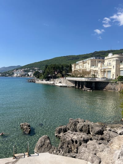 The image presents a picturesque coastal scene in Opatija, Croatia, captured on a clear, sunny day. The view looks along a tranquil bay, with a vibrant blue sky dotted with a few small white clouds occupying the upper left quadrant.
The main subjects include the clear, turquoise-blue waters of the sea, which gently ripple with reflected sunlight. On the right side, a rugged, dark gray rocky coastline forms the immediate edge, featuring concrete platforms and metal steps with handrails leading into the water, suggesting a public access point for swimming. A faint, distant figure appears to be swimming in the water near these steps.
Along the midground and extending into the background on the left, a lush green, tree-covered hillside slopes down to the water's edge. At the base of this hill, a series of buildings line the coast. Most prominent is a grand, multi-story, cream-colored edifice with ornate architectural details and numerous windows, likely a hotel or stately residence. The word "WAGNER" is clearly legible on the facade of this building. A white-balustraded promenade runs along the waterfront in front of the buildings, separating them from the sea. Further down the coast, more buildings of varying sizes create a charming townscape receding into the distance.
The scene depicts a calm and bright day, indicative of good weather for coastal activities. The clear water, bright sunlight, and architectural style suggest a popular resort or vacation destination. No specific activity beyond potential swimming or strolling along the promenade is clearly visible, but the overall impression is one of leisure and natural beauty.