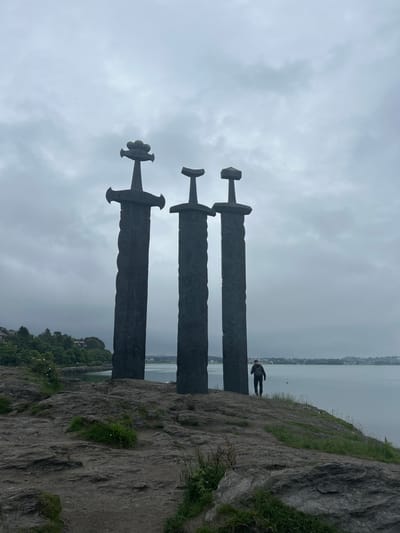 The image depicts the "Swords in a Rock" monument in Stavanger, Norway, a striking sculpture commemorating a historic Viking battle. Three colossal swords, each measuring 10 meters in height, are dramatically plunged into the rocky terrain. The swords are crafted from dark, textured metal, with intricate detailing on their hilts, suggesting an ancient, formidable design. The central sword is slightly taller than the other two.
The monument is situated on a clifftop overlooking a calm body of water, likely a fjord, reflecting the overcast sky. The surrounding landscape is rugged and natural, with scattered rocks, patches of green grass, and sparse vegetation. In the distance, across the water, a low-lying coastline with trees and buildings is visible, indicating the presence of a town or city.
A solitary figure, a man wearing a dark jacket and cap, stands near the base of the middle sword, facing the water. His posture suggests he is either observing the scenery or posing for a photograph. The man's presence provides a sense of scale, highlighting the immense size of the swords. The overall atmosphere of the image is somber and majestic, with the cloudy sky contributing to a dramatic and slightly melancholic mood. The lighting is diffused, creating soft shadows and a muted color palette dominated by grays, greens, and dark metallic tones.