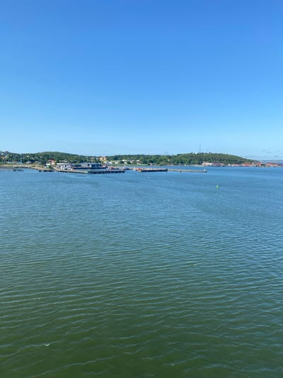 This media file depicts a coastal landscape under a clear, bright sky, likely taken during the daytime with good weather. The scene shows a body of water, possibly a harbor or bay, with a distant shoreline. The shoreline is lined with buildings, some appearing to be residential and others industrial or port-related. Green trees and vegetation are visible along the coast, suggesting a developed but also natural environment.

A long pier or breakwater extends into the water, with several boats or ships docked or moored nearby. One larger ship is prominently visible along the pier. There are also buoys in the water, including a bright green one, indicating navigational markers. The water itself is a murky green color and shows small ripples, suggesting a calm to moderately breezy condition.

The perspective seems to be from a slightly elevated viewpoint, possibly from a bridge, a boat, or a drone, looking out over the water towards the coast. The overall impression is one of a working port or coastal town, with a blend of industry and natural scenery. There are no visible people in the frame. The location context indicates this is likely Gothenburg, Sweden, which aligns with a coastal city environment featuring harbors and shipping activities.