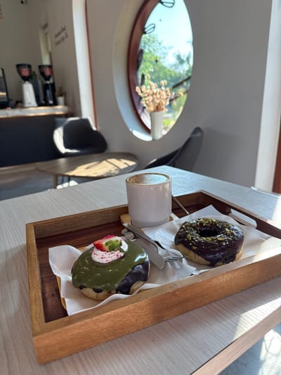 A forensic examination of the image reveals an indoor cafe setting, identified as located in San Phra Net, Thailand. The central focus is a wooden serving tray resting on a light-colored wooden table.
On the tray, from left to right:
1. A donut with dark green matcha glaze, topped with white cream, a half strawberry, and pink sprinkle-like particles, positioned on white parchment paper.
2. A white ceramic mug containing a light brown beverage with a frothy top layer, indicative of coffee or a latte.
3. A silver knife and fork, positioned between the mug and the second donut, resting on white parchment paper.
4. A donut with dark brown chocolate glaze, topped with crushed green nuts (pistachios), also positioned on white parchment paper.
The table exhibits a visible wood grain pattern, illuminated by natural light entering from the right, casting distinct shadows from the tray and its contents.
In the midground, the cafe interior is visible. To the left, a blurred counter area features at least two professional-grade coffee grinders and other cafe equipment. A dark grey upholstered chair is partially visible near a small, round wooden table. To the right, another dark grey chair is partially visible. A prominent oval-shaped window with a dark frame is visible on the right wall, through which natural light enters. On the window sill, a white cylindrical vase contains dried, light brown botanical material.
The background, viewed through the oval window, shows out-of-focus green foliage and a bright sky, indicating an outdoor environment during daylight hours. No individuals are present within the frame of the image.