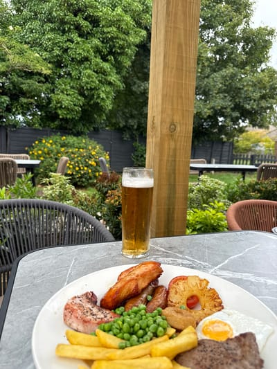 A close-up shot shows a plate of food and a glass of beer on a marble-patterned table. The plate contains a steak, fried egg, chips, peas, sausages, and grilled pineapple. In the background, a wooden post stands tall, with lush green trees and bushes behind it. A black wooden fence is visible in the distance, with some yellow flowers blooming in front of it. The scene appears to be set in an outdoor seating area of a restaurant or pub, possibly in High Wycombe, United Kingdom, given the typical setting of such establishments. The lighting suggests it is daytime, with an overcast sky contributing to the soft, natural illumination. There are no people visible in the frame.