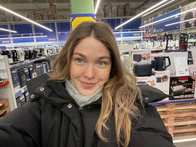 The image shows a person taking a selfie in what appears to be a large retail store, possibly a hypermarket or a warehouse club, as suggested by the visible "METRO" signage. The image is oriented upside down, with the person's head at the bottom.
The central subject is a young person, likely female, with fair skin, light-colored eyes (blue or grey), and long, wavy or slightly curly light brown hair with blonde highlights, which falls around her face and shoulders. She is wearing a grey hooded sweatshirt under a black puffer jacket with a high collar. Her expression is neutral, with a slight hint of a smile. Her lips are a natural pink color.
The background reveals rows of store shelves stocked with various products, predominantly kitchen appliances. To the person's left, an aisle features boxed items, including a "Tefal" brand toaster or grill with a price tag showing "14990" (possibly Russian Rubles, with a strikethrough price of "22990"). Below this, there are smaller black appliances, possibly kettles or coffee makers, from the "Moulinex" brand, packaged in white and red boxes. A black mug with a handle is visible in front of one of these boxes.
To the person's right, more shelves are filled with boxed goods, some appearing to be coffee makers or other small kitchen electronics from brands like "De'Longhi." The shelving units are metal, typical of a retail environment.
Further in the background, parallel to the aisles, there's a blue structure with white text that clearly reads "METRO" several times, indicating the name of the store. Below these blue signs, there are long, linear fluorescent lights running across the ceiling, illuminating the store. On the left side of the image, another blue sign with white text is visible, which appears to read "КАССЫ" (KASSY), meaning "Checkouts" or "Cashiers" in Russian. The floor is light-colored.
On the far left, partially obscured, is a stack of wooden pallets, suggesting goods are stored or displayed in a warehouse-style setting. The overall impression is a casual, everyday shopping trip in a large, well-lit retail space.