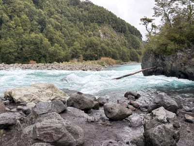The image captures a river landscape in Puerto Varas, Chile. The water is a vibrant turquoise, flowing rapidly over rocks and creating white water. A fallen log spans the river, partially submerged. Large grey boulders are scattered along the riverbank in the foreground, and a steep hillside covered in dense green trees rises on one side of the river. The sky is overcast. No people are visible.