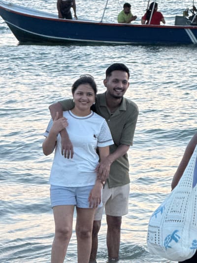 The image depicts a scene at Cenang Beach, Langkawi, Malaysia, featuring individuals in shallow water with a boat in the background. In the foreground, a man and a woman stand in knee-deep, rippled water, both smiling and looking forward. The woman, positioned on the left, wears a white short-sleeved t-shirt with grey striped details on the sleeves and collar, paired with light blue shorts. Her right hand rests on the man's left arm. The man, positioned behind and slightly to the right of the woman, wears an olive green short-sleeved collared shirt and light-colored shorts. His right arm is around the woman's upper back, and his left hand is placed near her waist. To the far right, a partially visible individual holds a white crocheted bag adorned with light blue turtle designs. In the mid-ground, a blue and white boat floats on the water. Three individuals are visible within the boat: one person in a dark top standing near the bow, one person in a bright green top seated, and another person in a red top also seated. The ambient lighting suggests a late afternoon or early evening setting.