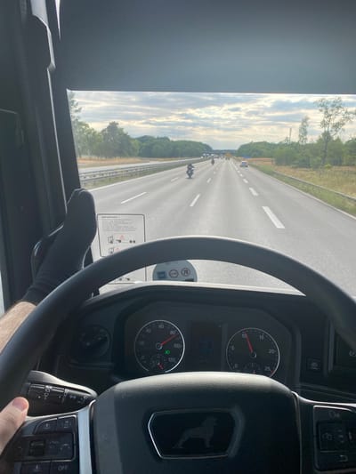 The image is taken from the driver's perspective inside a truck, looking through the windshield at a highway in Ludwigsfelde, Germany. The truck is traveling on a multi-lane highway with moderate traffic, including several motorcycles and cars. The sky is cloudy, with sunlight breaking through. On the dashboard, the steering wheel is visible, along with various controls and a speedometer displaying approximately 70 km/h. A pair of black socks is visible on the driver's left foot, which is resting on or near the dashboard. A sticker with important driving information is attached to the window pillar. The environment outside the truck consists of green trees and vegetation lining the highway.