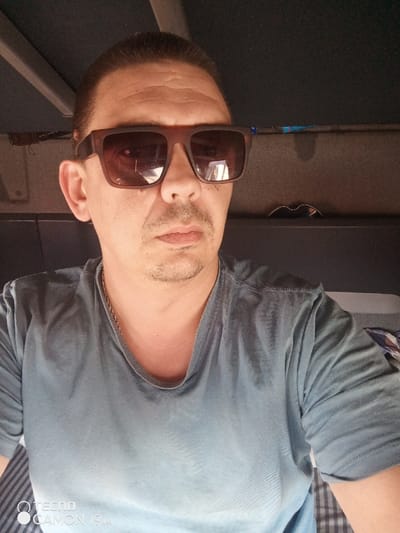 A close-up photograph features a male individual positioned in an interior setting, identified as being in Pridorozhnyi, Russia. The male has short, dark hair slicked back, a mustache, and a goatee. He wears dark brown, thick-framed sunglasses and a V-neck t-shirt, which is dark blue-grey at the top and fades to a lighter blue-grey towards the bottom. A thin, gold-colored chain is present around his neck. His expression is neutral as he faces the camera directly. The background suggests the cabin of a vehicle, with a dark ceiling visible overhead and a light-colored structural beam on the upper left. A dark shelf to the right holds a pair of dark-rimmed glasses. Below this, a section of horizontally striped fabric, appearing as dark grey and light grey, is partially visible. The bottom left corner of the image displays a "TECNO CAMON 19 Pro" watermark.