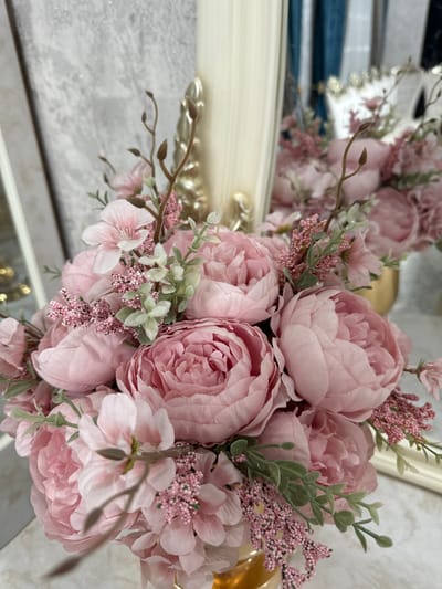 A bouquet of light pink peonies and smaller blossoms fills the frame, accented with small green leaves and brown stems. Some flowers are fully open, while others are still tightly budded. A reflection of the floral arrangement is visible in a shiny surface to the left, with hints of blue and white in the background. The lighting highlights the soft textures and delicate colors of the flowers.