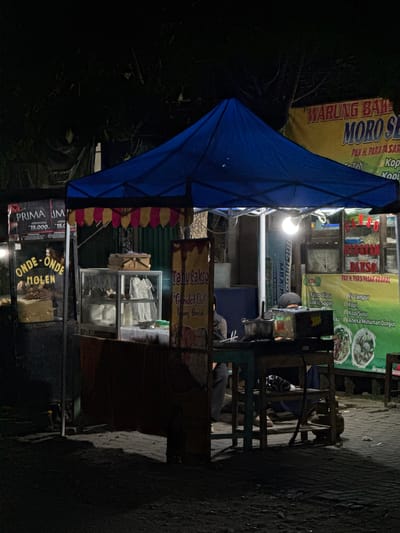 The image depicts a nighttime scene at a food stall in Alian, Indonesia. A blue canopy tent is set up, illuminated by a bright overhead light. Under the tent, there are various food displays and cooking equipment. To the left, a sign for "PRIMA IM" and "ONDE-ONDE MOLEN" is visible. A glass display case contains food items, and a person is seen working behind it. To the right, a banner for "WARUNG BAK MORO SE" is displayed, listing menu items like "Bakso", "Es Campur", "Kopi", and "Aneka Minuman Dingin". Another banner shows images of food. Several people are present, including one person wearing a dark head covering and another partially visible person in the background. The ground is paved with stones, and shadows are cast by the lighting. A gas cylinder is visible near a wooden table. The overall impression is of a busy street food stall operating at night.