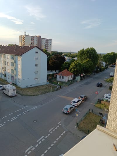 This media file captures an outdoor scene in Schwetzingen, Germany, during what appears to be late afternoon or early evening, with the sun casting long shadows. The sky is partly cloudy, with a clear blue expanse above.

The foreground is dominated by a residential building with a light blue facade and a tiled roof. An RV is parked near the building. Across the street, a paved road is visible with several cars parked along it and in motion. Among the vehicles are a white RV, a dark sedan, and several passenger cars. A small, single-story building with a red tile roof is situated further down the street, surrounded by greenery. In the background, two tall apartment buildings stand out, with several smaller houses and trees interspersed throughout the landscape. Power lines and transmission towers are faintly visible in the distance.

Several street signs are present, including one with the number "30" indicating a speed limit, and a triangular warning sign with a red border. The street is marked with white dashed lines, suggesting a road intersection. A bicycle is parked near a small fenced-off grassy area. A lamppost stands at the edge of the grass.

The scene depicts a quiet, everyday moment in a residential neighborhood, with no specific event or activity clearly taking place beyond typical traffic and parked vehicles. The overall mood is calm and tranquil, bathed in the soft light of the setting sun. The text "Karlstraße 52-54" is visible on the side of the blue residential building.