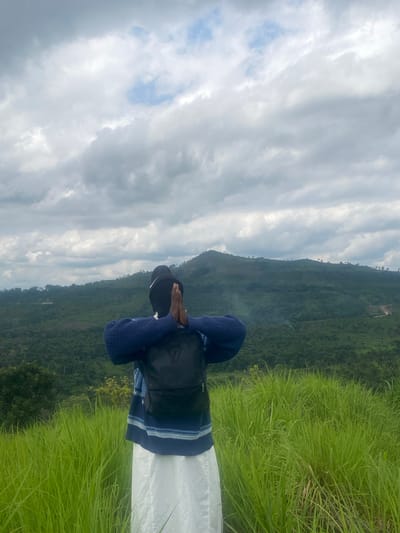 The image depicts a person standing on a grassy hill, facing away from the camera and looking out at a lush, green landscape. The person is wearing a blue and white striped sweater over a black backpack and a white skirt. Their hands are pressed together in front of their face, suggesting a moment of reflection or prayer. The background features rolling hills covered in dense vegetation, with a cloudy sky overhead. The scene appears to be a natural outdoor setting, likely in a rural area, and given the user's prompt, this could be the city of Unwana, Nigeria. The lighting is overcast, indicating a cloudy day.