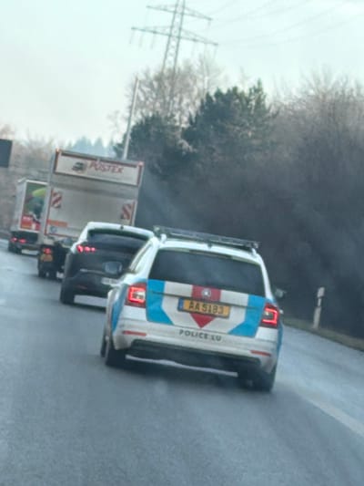 This image depicts a line of vehicles on a wet asphalt road in Itzig, Luxembourg, under an overcast sky. In the foreground, the rear of a white and light-blue Luxembourgish police station wagon is visible, angled slightly left, displaying license plate "AA 5193" and "POLICE.LU" below it. Its brake lights are off. Directly ahead of the police vehicle is a dark-colored passenger car. Further ahead, a white trailer truck with "POSTEX" emblazoned on its rear is followed by another truck, indicating queued traffic. To the right of the road, a dense line of trees, including both deciduous and evergreen varieties, borders the scene. A large electricity transmission tower stands against the light grey sky in the upper background, positioned above the tree line. The road surface appears wet, suggesting recent precipitation.