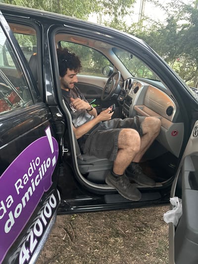 A man is contorted inside the driver's side of a car, seemingly resting or lounging, with his legs propped up on the passenger side. He is holding a cell phone in his hand. The car door is open, revealing the vehicle's interior. A sticker with the words "Radio a Domicilio" and the number "420100" is on the car. The car is parked outdoors with greenery visible in the background.