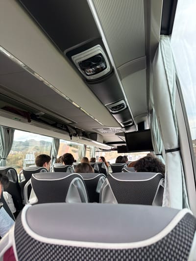 The image presents an interior view of a passenger bus in Zarautz, Spain, captured from a seated position looking forward. Multiple individuals, primarily young adults, occupy the bus's grey and black patterned seats, facing the front of the vehicle. Their backs and heads are visible. The bus features a light grey ceiling with dark grey central panels containing integrated overhead lighting units and air vents. On the left, an open overhead storage rack holds several items, including what appear to be umbrellas. Light-colored curtains are pulled back and secured on both the left and right side windows. Through the windows, a hilly urban landscape is visible, featuring buildings and a road or bridge structure under an overcast sky. A dark, blank flat-screen monitor is mounted from the ceiling on the right side of the aisle.