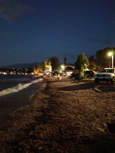 The image shows a beachside area at dusk or night, likely in the Municipal Unit of Vrachneika, Greece. The foreground is a sandy or gravelly beach with some vegetation. Several vehicles are parked on the beach, including a white SUV in the right foreground. In the background, there is a body of water with gentle waves breaking on the shore. Beyond the water, a line of lights suggests a town or settlement along the coast. Trees and a streetlamp are visible, contributing to the evening ambiance. The sky is dark blue, indicating it is either twilight or nighttime. There are no people clearly visible in the frame.