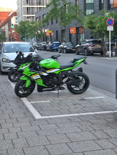 This detailed image captures an urban street scene in Sanem, Luxembourg, featuring vehicles and modern architecture under a soft, possibly late afternoon, light.
In the immediate foreground, a vibrant green, black, and white Kawasaki Ninja ZX-6R motorcycle is parked on a light grey paver sidewalk, positioned within a clearly marked white rectangular parking space designated for motorcycles. The motorcycle is on its kickstand and faces towards the left side of the frame. Partially visible directly behind the green motorcycle is the front section of a black motorcycle, also parked on the sidewalk.
On the asphalt roadway adjacent to the sidewalk, a silver-white Volkswagen hatchback is visible in the foreground, facing away from the viewer. Further down the street, a line of cars is parked or stationary. These include a black sedan, a dark grey SUV with a yellow rear license plate, and a blue sedan, all featuring yellow European-style license plates characteristic of Luxembourg.
The street is flanked by modern multi-story buildings. On the right, the buildings have dark grey or charcoal facades with numerous windows. On the left, the buildings appear lighter in color, possibly white or light grey, with varied window structures. Green-leafed trees are planted intermittently along the sidewalks on both sides of the street.
Environmental details include a red circular "No Stopping" sign (R 205) mounted on a pole on the right side of the street, with an unreadable white rectangular sign beneath it. A green traffic light is visible on the left side of the street in the distance. No individuals are discernible within the frame.