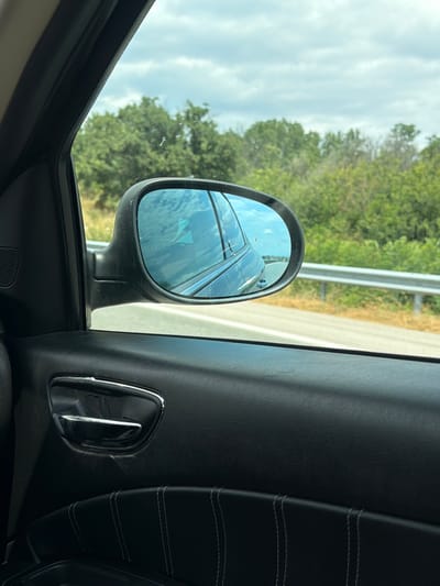 The image shows the interior of a car, looking out the passenger side window. A side-view mirror reflects the sky and trees. The car appears to be driving on a road lined with a metal guardrail and dry grass. The interior of the car is dark, with black leather trim and a chrome door handle visible. The location is identified as Aleksandrovo, Bulgaria.