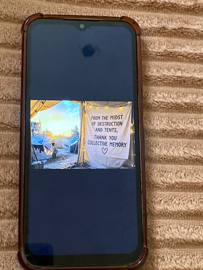 A smartphone, encased in a dark brown protective cover, is shown resting on a textured, light brown surface, possibly a blanket or towel. The phone screen displays an image that is rotated 90 degrees counter-clockwise. There's a dark smudge on the upper left side of the phone's screen, near the camera notch.

The image displayed on the phone consists of two distinct parts, vertically stacked when viewed in its intended orientation. The upper section features a white banner or cloth hung up, adorned with string lights. On this banner, dark text is written, which reads: "FROM THE MIDST OF DESTRUCTION AND TENTS, THANK YOU COLLECTIVE MEMORY ♡". A small heart symbol is visible at the end of the text.

The lower section of the displayed image depicts an outdoor scene at what appears to be either sunrise or sunset, indicated by the warm, bright light radiating from the horizon. The foreground shows several makeshift structures or tents within what looks like a camp or a damaged settlement. The ground appears uneven, possibly covered in rubble or dirt. Silhouettes of distant structures are visible against the bright sky. Given the context of al-Qarara, Palestinian Territories, this scene likely portrays a temporary settlement or an area affected by conflict or displacement. The combination of destruction, tents, and the message of "collective memory" conveys a sense of hardship endured by a community, accompanied by gratitude and remembrance.