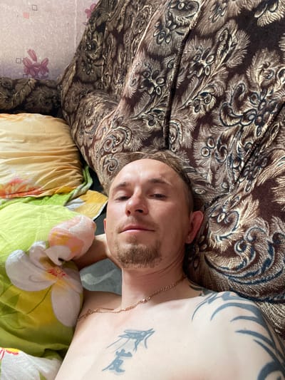 A male individual is lying on a bed in a room. He has light skin, short blond hair, a goatee, and is wearing a gold chain. He has tattoos on his upper left arm and chest. The bed is covered with a patterned brown fabric, and there are pillows with floral patterns. The background shows a wall with a floral wallpaper. The scene is shot from a high-angle perspective. The location is identified as Nizhnii Bugrysh, Russia.